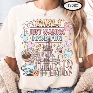 May include: Ivory-colored t-shirt with the text "GIRLS JUST WANNA HAVE FUN" in pastel colors, along with cartoon illustrations of a castle, Mickey Mouse, popcorn, and other magical elements.
