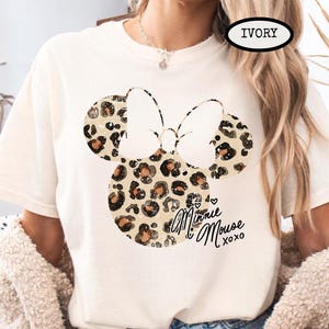 May include: Ivory t-shirt with a Minnie Mouse design. The design features a bow and mouse ears with a leopard print pattern. The words "Minnie Mouse xoxo" are written in script below the design.