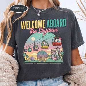 May include: Dark gray t-shirt with a colorful graphic of a gondola ride with the text "WELCOME ABOARD The Skyliner". The design includes cartoon characters, a sphere, and a castle.