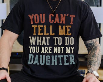 May include: Black t-shirt with the text "YOU CAN'T TELL ME WHAT TO DO YOU ARE NOT MY DAUGHTER" in a retro font. The shirt is a dark color and has a vintage look.