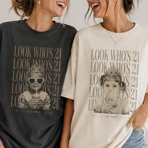 May include: Two oversized crewneck t-shirts, one charcoal gray and one cream, each featuring a graphic print of a child wearing a crown and heart-shaped sunglasses. The text "LOOK WHO'S 21" is repeated vertically above the image, with "EST. 2003" below.