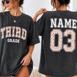 May include: A charcoal gray short-sleeve t-shirt featuring a floral pattern inside the letters of the word THIRD and the number 03. The front displays the text THIRD GRADE, while the back shows the word NAME above the number 03.
