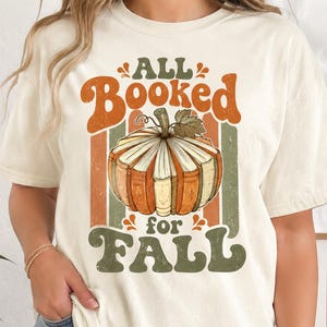 May include: A cream-colored short-sleeve t-shirt featuring a graphic of a pumpkin made of stacked books. The text reads "ALL BOOKED for FALL" in orange and green retro-style lettering, set against vertical stripes of orange, green, and cream.