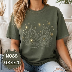 May include: A moss green short-sleeve t-shirt featuring a white line art illustration of wildflowers and grasses. The design includes glowing fireflies scattered among the plants and the text Summer Nights printed in a cursive script font.