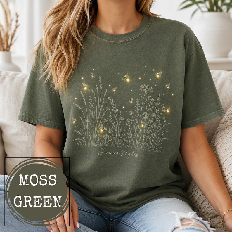 May include: A moss green short-sleeve t-shirt featuring a white line art illustration of wildflowers and grasses. The design includes glowing fireflies scattered among the plants and the text Summer Nights printed in a cursive script font.