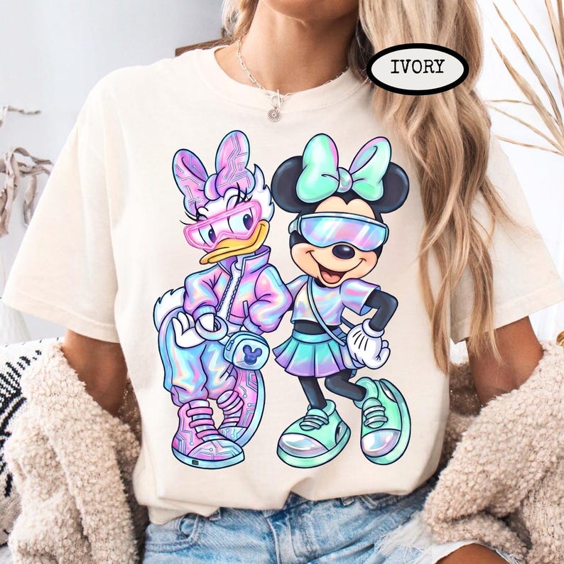 May include: Ivory-colored t-shirt featuring a colorful graphic of Daisy Duck and Minnie Mouse in futuristic outfits. Daisy Duck wears pink and blue, while Minnie Mouse sports a mint green and purple ensemble. The shirt has a crew neck and short sleeves.