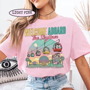 May include: Light pink t-shirt with a colorful graphic featuring the text "WELCOME ABOARD The Skyliner." The design includes gondolas, a moon, palm trees, and architectural landmarks. The shirt is a crew neck style.