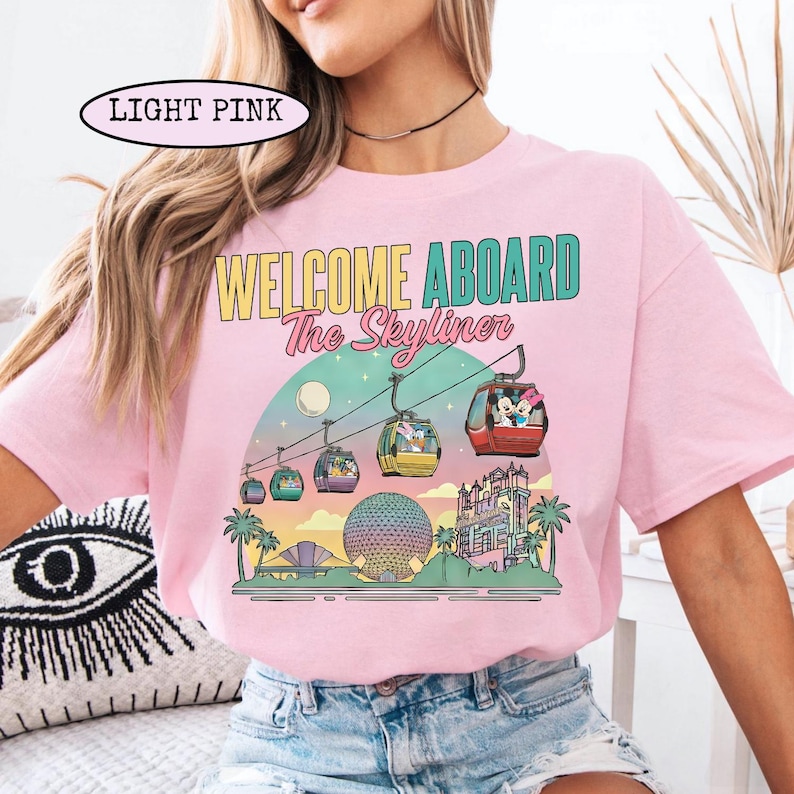 May include: Light pink t-shirt with a colorful graphic featuring the text "WELCOME ABOARD The Skyliner." The design includes gondolas, a moon, palm trees, and architectural landmarks. The shirt is a crew neck style.