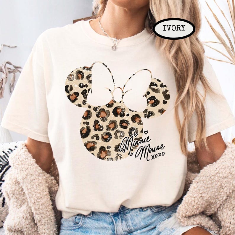 May include: Ivory t-shirt with a Minnie Mouse design. The design features a bow and mouse ears with a leopard print pattern. The words "Minnie Mouse xoxo" are written in script below the design.
