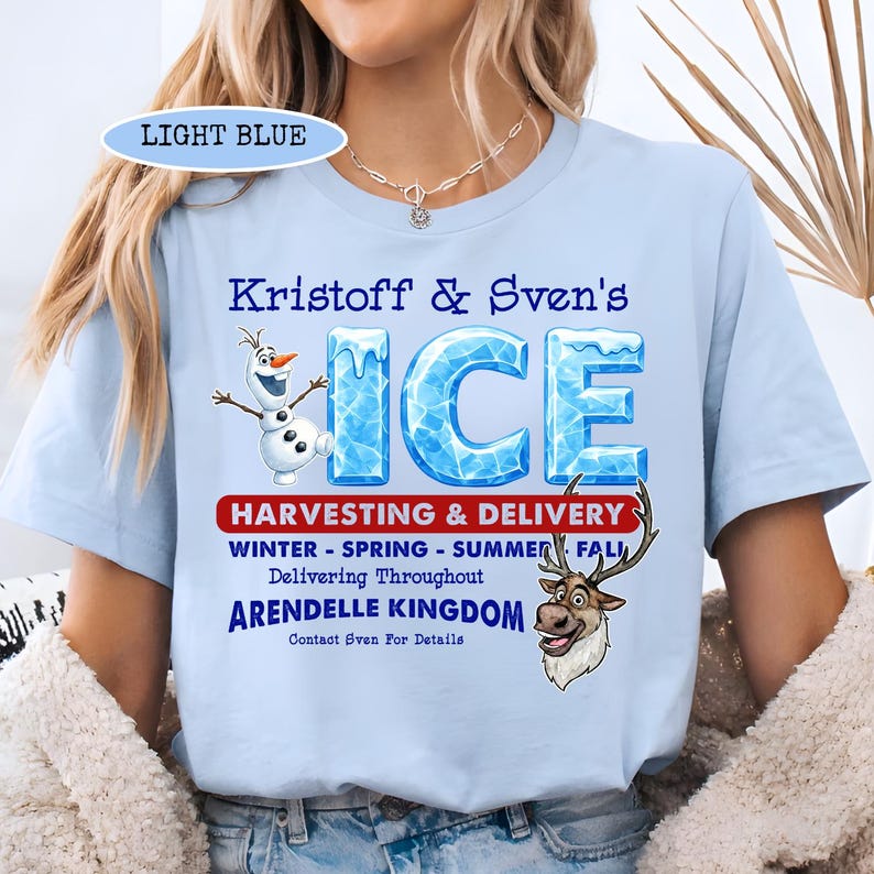 Op de afbeelding: Lichtblauw T-shirt met de tekst "Kristoff & Sven's ICE" en afbeeldingen van Olaf en Sven. Het shirt bevat ook tekst over het oogsten en bezorgen van ijs in het koninkrijk Arendelle.