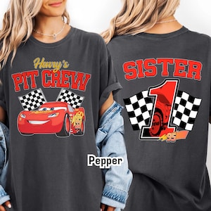 May include: Dark gray t-shirt featuring a red race car graphic with the text "Henry's Pit Crew" on the front. The back of the shirt says "Sister 1" with a race car graphic and checkered flags.