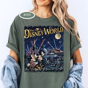 May include: Moss green t-shirt featuring a colorful graphic of Walt Disney World at night. The design includes Mickey and Minnie Mouse, fireworks, and the text "Walt Disney World" and "The Happiest Place on Earth."