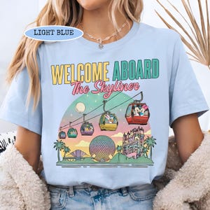 May include: Light blue t-shirt with the text "WELCOME ABOARD The Skyliner" and a graphic of a gondola ride with cartoon characters. The design includes a sunset, palm trees, and a large sphere.