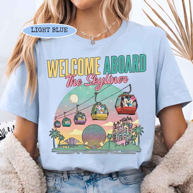 May include: Light blue t-shirt with the text "WELCOME ABOARD The Skyliner" and a graphic of a gondola ride with cartoon characters. The design includes a sunset, palm trees, and a large sphere.