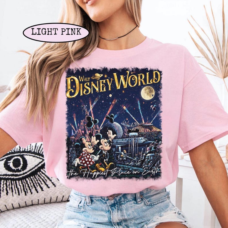 May include: Light pink t-shirt featuring a Walt Disney World graphic. The design includes Mickey and Minnie Mouse, fireworks, and the text "The Happiest Place on Earth." The shirt is a soft, comfortable material.