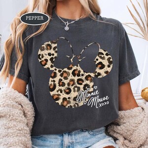 May include: Dark grey t-shirt with a leopard print Minnie Mouse design. The design includes the words "Minnie Mouse xoxo" in a cursive font. The shirt has a crew neck and short sleeves.