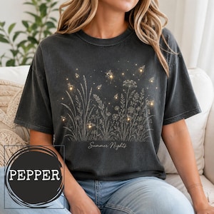 May include: A charcoal gray short-sleeve t-shirt featuring a white line art illustration of wildflowers and tall grasses. The design includes small glowing yellow dots representing fireflies and the text Summer Nights in script font.