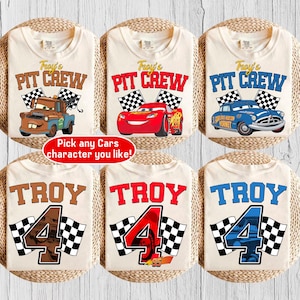May include: Six beige t-shirts with racing-themed designs. The shirts feature the text "Troy's Pit Crew" and "Troy 4" with checkered flags and cartoon car graphics. One shirt has the text "Pick any Cars character you like!"