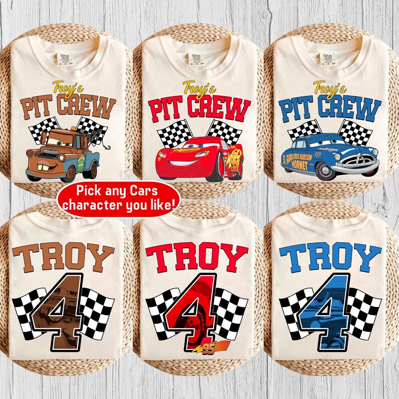 May include: Six beige t-shirts with racing-themed designs. The shirts feature the text "Troy's Pit Crew" and "Troy 4" with checkered flags and cartoon car graphics. One shirt has the text "Pick any Cars character you like!"