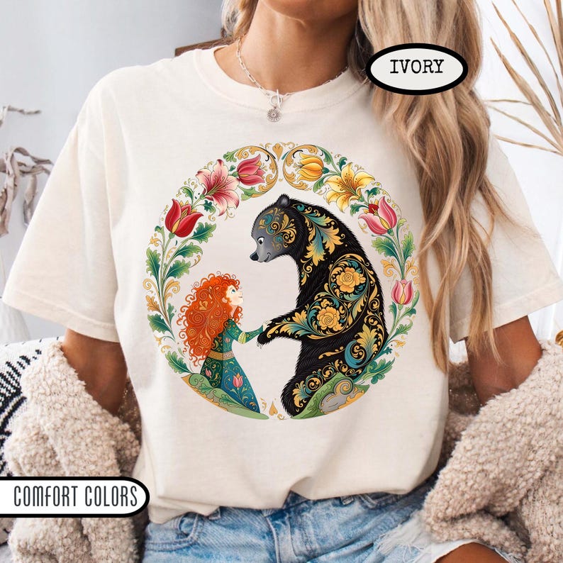 May include: Ivory-colored t-shirt with a circular floral design featuring a black bear and a red-haired figure. The bear has ornate gold and teal patterns. The text "IVORY" and "COMFORT COLORS" are printed on the shirt.