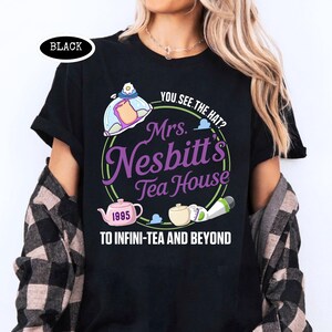 Puede incluir: Camiseta negra con un dise&ntilde;o gr&aacute;fico que presenta el texto "Mrs. Nesbitt's Tea House" en morado y verde, junto con la frase "To Infini-Tea and Beyond". El dise&ntilde;o incluye una tetera y otras ilustraciones relacionadas con el t&eacute;.