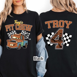 May include: Black t-shirt featuring a cartoon tow truck with the text "Troy's Pit Crew" and checkered flags. The back of the shirt displays "TROY" and the number "4" with checkered flags. The shirt is black.