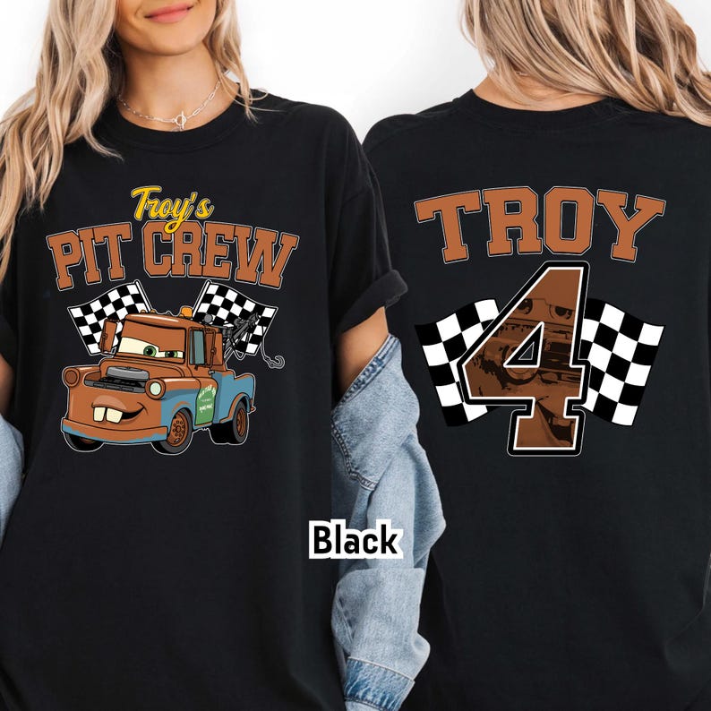 May include: Black t-shirt featuring a cartoon tow truck with the text "Troy's Pit Crew" and checkered flags. The back of the shirt displays "TROY" and the number "4" with checkered flags. The shirt is black.