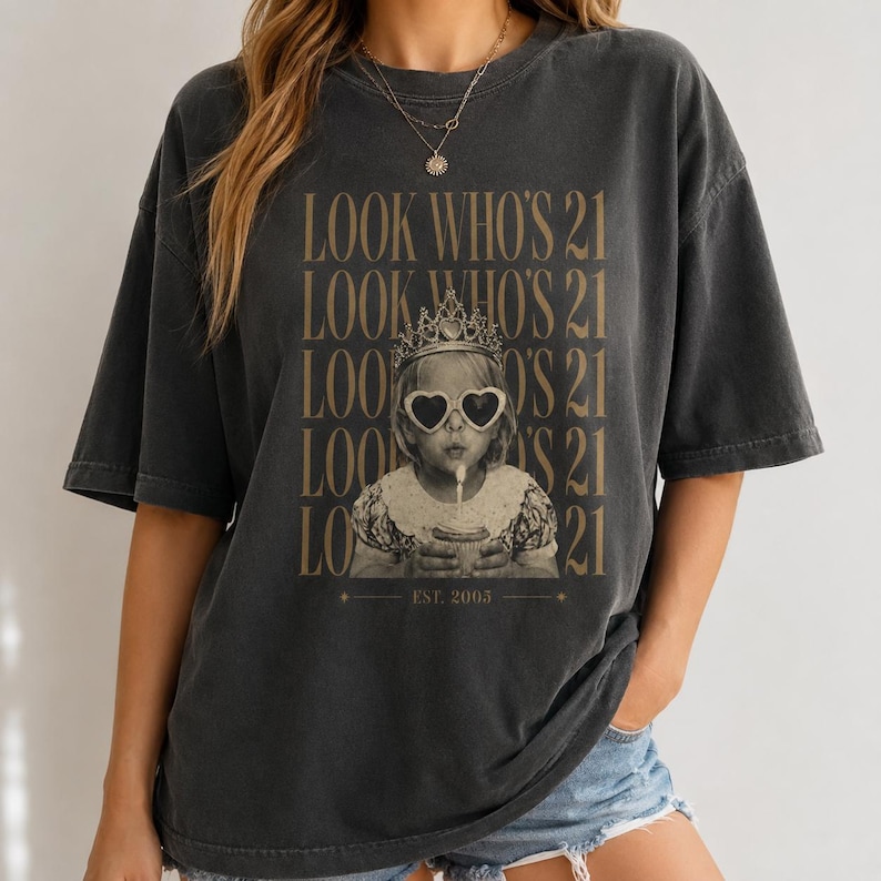 May include: A charcoal gray short-sleeve t-shirt featuring a graphic print of a child wearing a crown and heart-shaped sunglasses while holding a cupcake. The text "LOOK WHO'S 21" is repeated five times in tan, with "EST. 2005" printed at the bottom.