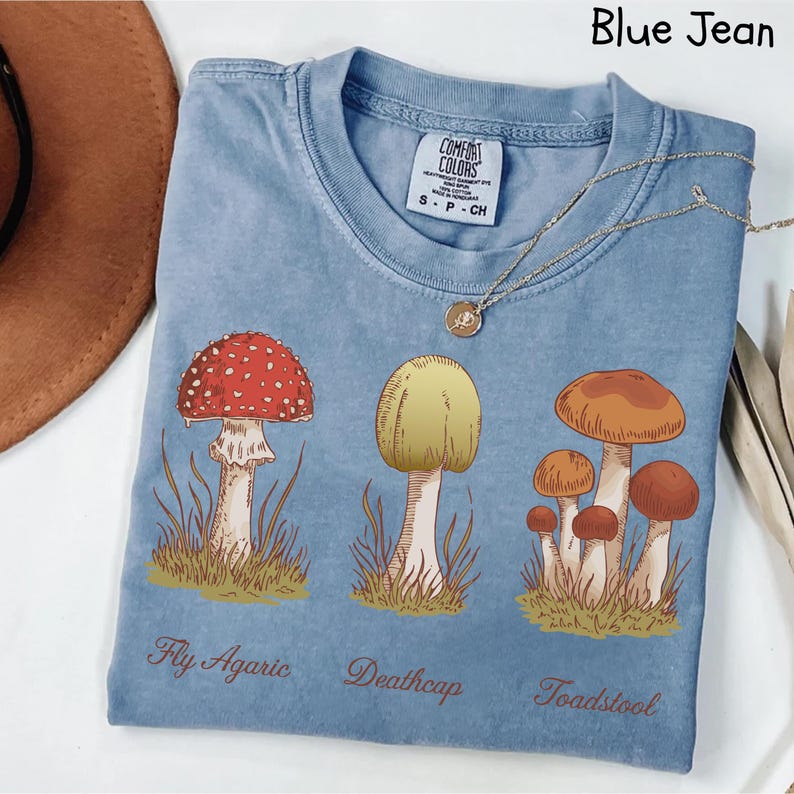 May include: A blue jean t-shirt with a mushroom graphic. The shirt features three different types of mushrooms: Fly Agaric, Deathcap, and Toadstool. The shirt has the text "Comfort Colors" on the tag.