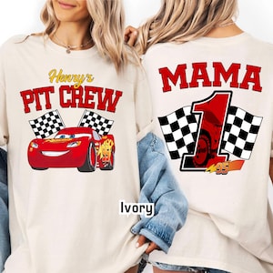 May include: Ivory-colored t-shirts with racing-themed graphics. One shirt features "Henry's Pit Crew" with a red race car and checkered flags. The other shirt displays "MAMA 1" with a race car and checkered flags. The number "95" is also visible.