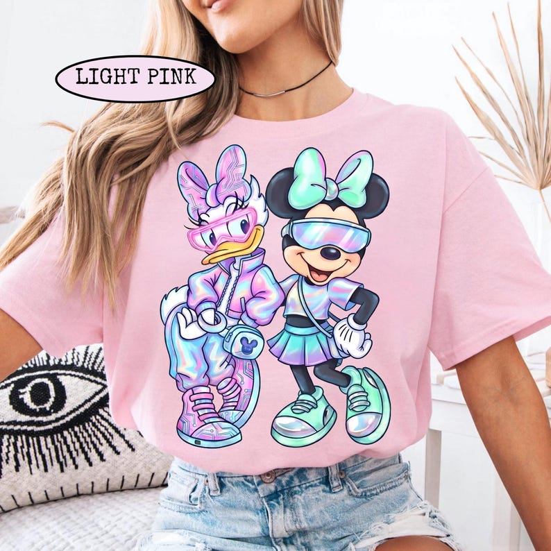May include: Light pink t-shirt featuring a graphic of Daisy Duck and Minnie Mouse wearing futuristic outfits and sunglasses. The text "LIGHT PINK" is above the graphic. The characters are in a holographic color scheme with mint green and light blue accents.