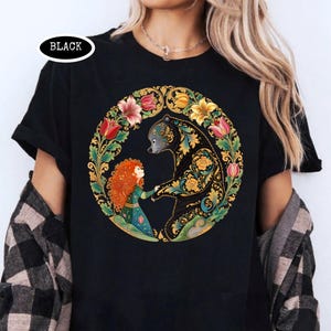 May include: Black t-shirt featuring a circular design with a bear and a girl holding hands, surrounded by flowers. The bear is black with gold and teal patterns. The girl has red hair and wears a green dress. The word "BLACK" is in a white oval.