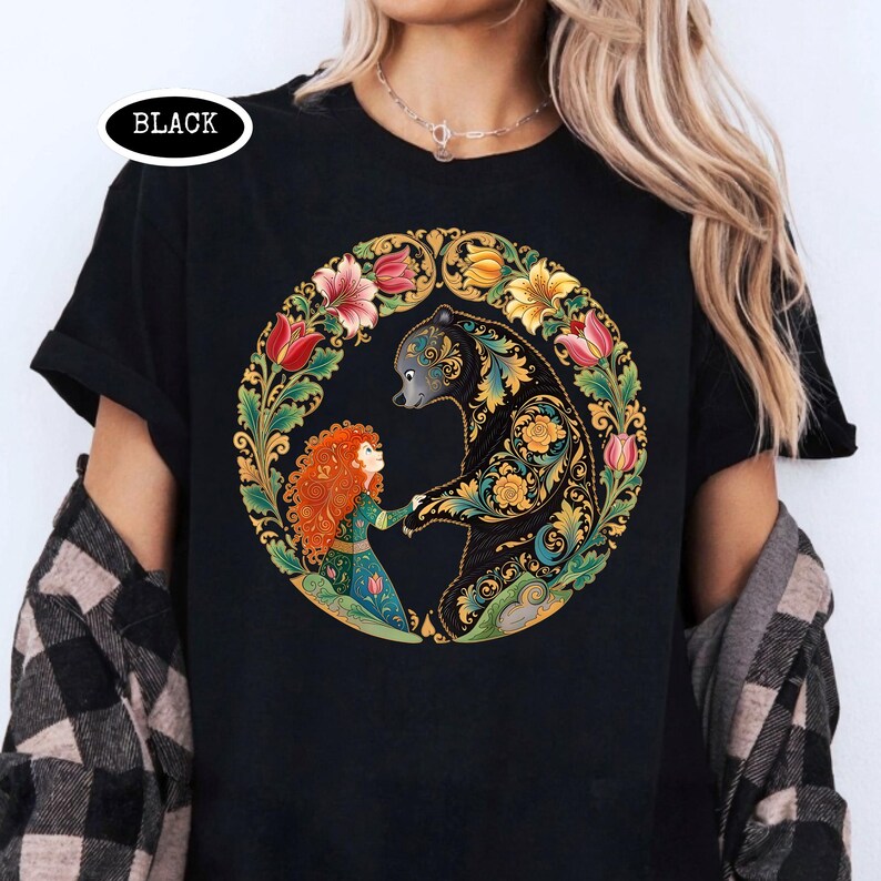 May include: Black t-shirt featuring a circular design with a bear and a girl holding hands, surrounded by flowers. The bear is black with gold and teal patterns. The girl has red hair and wears a green dress. The word "BLACK" is in a white oval.