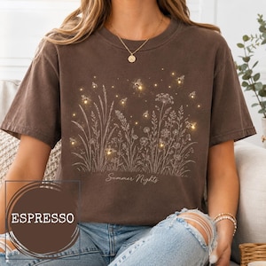 May include: A dark brown short-sleeve t-shirt featuring a white line-art illustration of wildflowers and fireflies. The text Summer Nights is printed below the graphic. A circular overlay in the corner displays the word ESPRESSO in white text.
