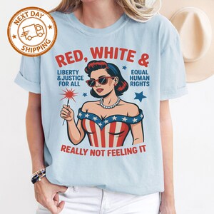 May include: Light blue t-shirt with a graphic of a woman in sunglasses and a patriotic outfit, holding a sparkler. Text on the shirt reads "RED, WHITE & EQUAL HUMAN RIGHTS, LIBERTY & JUSTICE FOR ALL, REALLY NOT FEELING IT."