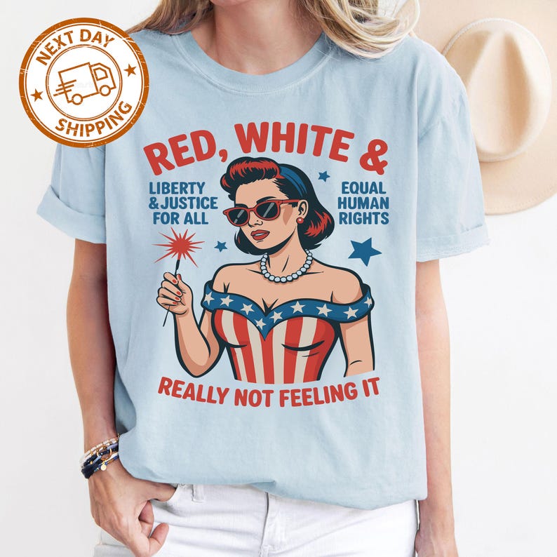 May include: Light blue t-shirt with a graphic of a woman in sunglasses and a patriotic outfit, holding a sparkler. Text on the shirt reads "RED, WHITE & EQUAL HUMAN RIGHTS, LIBERTY & JUSTICE FOR ALL, REALLY NOT FEELING IT."