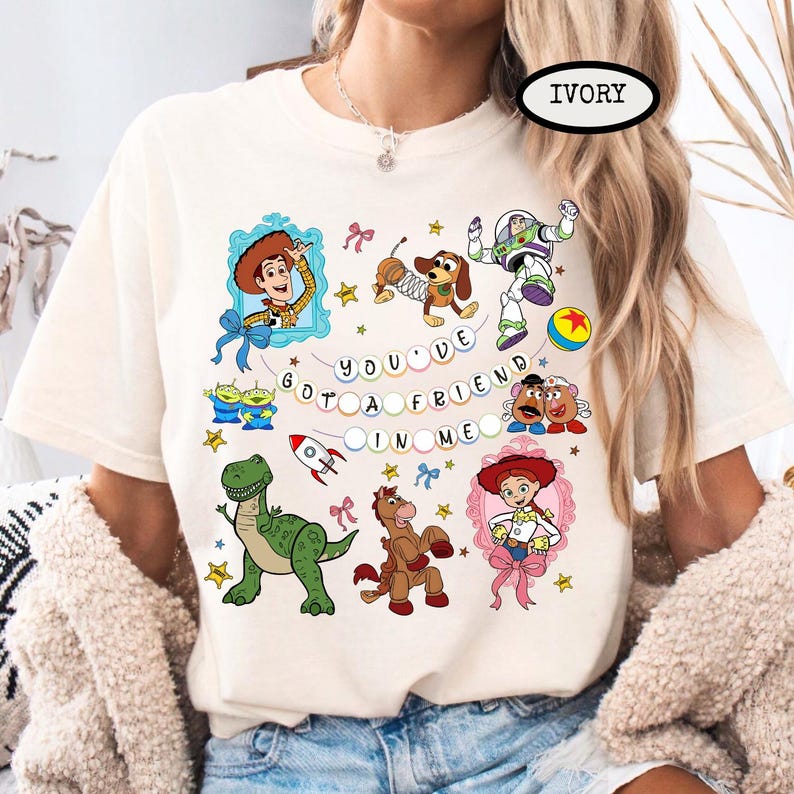 May include: Ivory-colored t-shirt featuring colorful cartoon characters and the text "YOU'VE GOT A FRIEND IN ME." The design includes images of Woody, Buzz Lightyear, and other characters from the animated movie.
