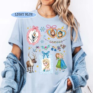 Puede incluir: Camiseta azul claro con varios personajes y elementos de la pel&iacute;cula animada "Frozen". El dise&ntilde;o incluye im&aacute;genes de Olaf, Anna, Elsa, Kristoff y Sven, junto con la palabra "FROZEN" y marcos decorativos y copos de nieve.