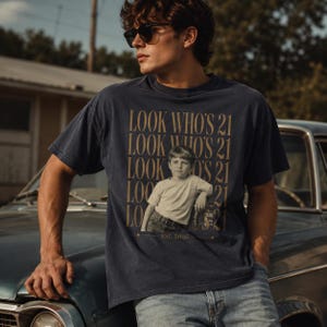 May include: A dark navy blue short-sleeve t-shirt featuring a graphic print of a young child in a light-colored shirt and jeans. The text "LOOK WHO'S 21" is repeated in gold lettering, with "EST. 2005" printed at the bottom of the graphic design.