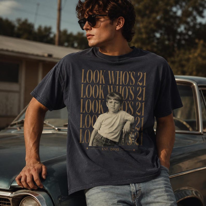 May include: A dark navy blue short-sleeve t-shirt featuring a graphic print of a young child in a light-colored shirt and jeans. The text "LOOK WHO'S 21" is repeated in gold lettering, with "EST. 2005" printed at the bottom of the graphic design.