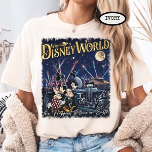 May include: Ivory-colored t-shirt featuring a colorful graphic of Walt Disney World at night, with Mickey and Minnie Mouse. The text "Walt Disney World" and "the Happiest Place on Earth" are also displayed.