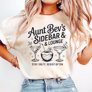 May include: Cream-colored t-shirt with the text "Aunt Bev's Sidebar & Lounge" in black script. Below the text are three cocktail illustrations. The phrase "Stay Salty, Resist Often" is printed below the illustrations.