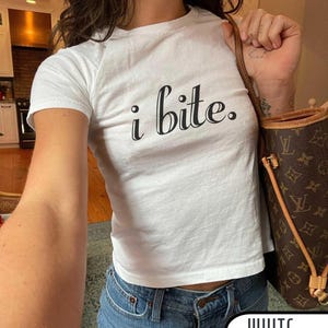 May include: White crop top with the text "i bite." in black script. The top is paired with blue jeans. A brown handbag is visible in the image. The word "WHITE" is written in a white box at the bottom of the image.