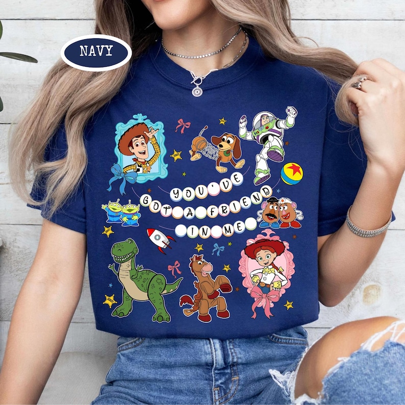 May include: Navy blue t-shirt featuring colorful cartoon characters and the text "YOU'VE GOT A FRIEND IN ME." The design includes Woody, Buzz Lightyear, Rex, and other characters from the animated movie. The shirt has a crew neck.