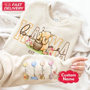 Könnte beinhalten: Cremefarbenes Sweatshirt mit Winnie-the-Pooh-Design. Das Design zeigt das Wort "Mama", Winnie Puuh mit einem Honigtopf, Blumen und Bienen. Die Ärmel haben Ballons mit Charakterillustrationen und individuellen Namen.