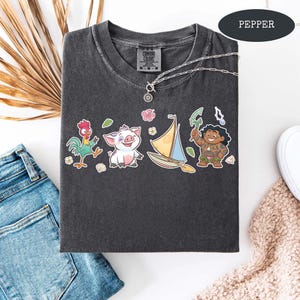 Op de afbeelding: Een donkergrijs T-shirt met een ronde hals en een kleurrijke cartoonafbeelding. Het shirt heeft een zilveren ketting met een hanger. Het woord "PEPPER" staat in een donker ovaal rechtsboven.