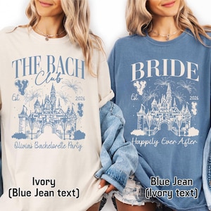 May include: Two t-shirts featuring a castle graphic with fireworks and balloons. The ivory shirt displays 'THE BACH Club' and 'Olivia's Bachelorette Party' in blue text. The blue jean shirt displays 'BRIDE' and 'Happily Ever After' in ivory text.