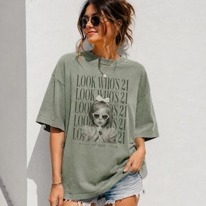 May include: An olive green short-sleeve t-shirt featuring a repeating black text graphic that reads LOOK WHO'S 21. The center graphic displays a black and white photograph of a young child wearing heart-shaped sunglasses and a bow, with EST. 2003.