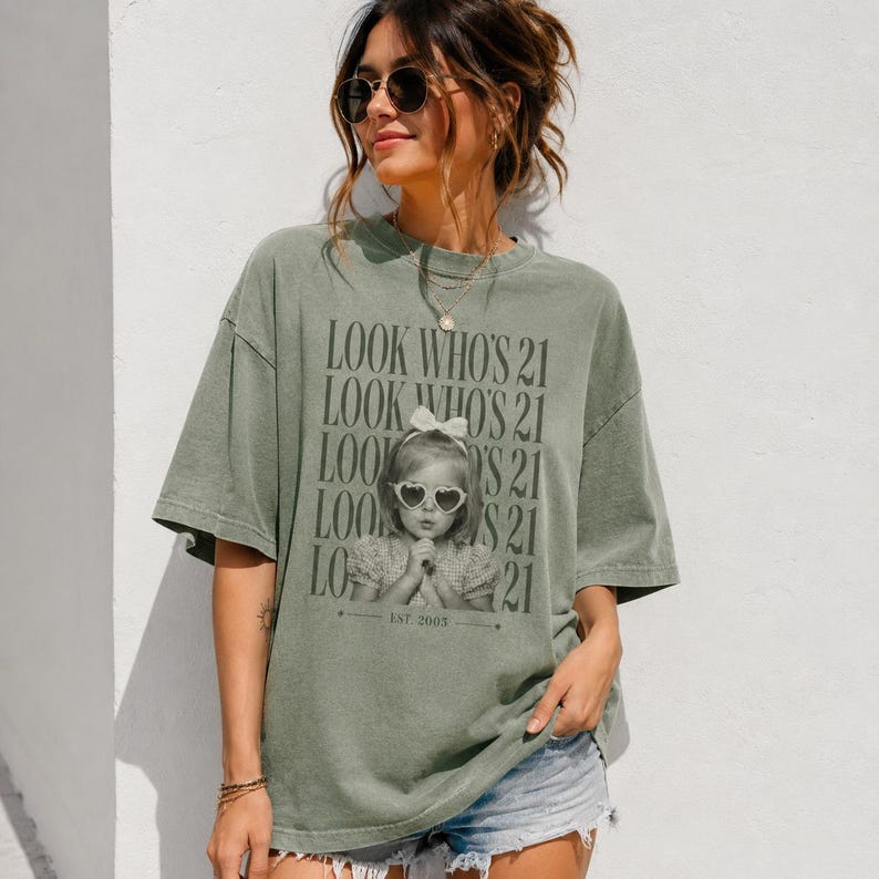 May include: An olive green short-sleeve t-shirt featuring a repeating black text graphic that reads LOOK WHO'S 21. The center graphic displays a black and white photograph of a young child wearing heart-shaped sunglasses and a bow, with EST. 2003.
