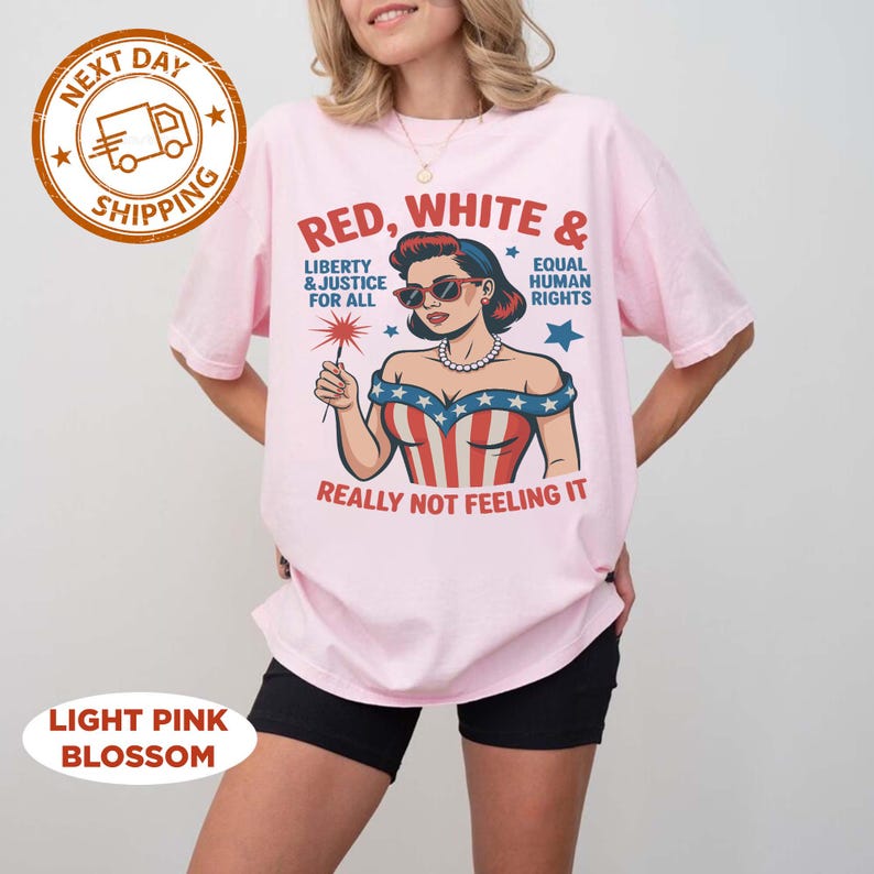 May include: Light pink t-shirt with a graphic of a woman in sunglasses holding a sparkler, with text that reads "Red, White & Liberty & Justice For All, Equal Human Rights, Really Not Feeling It".